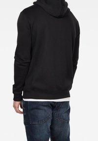 Felpa nera con cappuccio, caratterizzata da una texture semplice e liscia, con polsini e orlo a costine, indossata sopra una camicia bianca, abbinata a jeans blu.