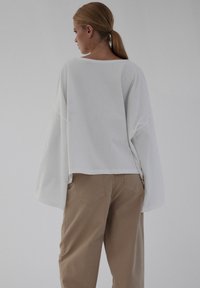 Biała, luźna bluzka z szerokimi rękawami, zestawiona z jasnobrązowymi, luźno dopasowanymi spodniami z tylnymi kieszeniami i gładką teksturą.