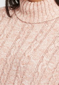 Kabelstickad polotröja i mjuk rosa och krämfärgad garn, med en chunky textur och ribbad krage, lämplig för avslappnad användning.