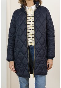Una persona indossa un cappotto trapuntato blu navy sopra un maglione a righe e jeans, in piedi contro uno sfondo chiaro. Il cappotto ha una cerniera dorata.
