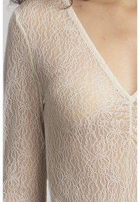 Close-up van een persoon die een doorschijnende, beige langslavige top draagt met ingewikkelde bloemige kantpatronen en een V-hals.