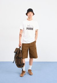 Mann i svart bøttehatt, hvit Puma t-skjorte, brune shorts og joggesko, som holder en brun ryggsekk i et hvitt rom med blått gulv.