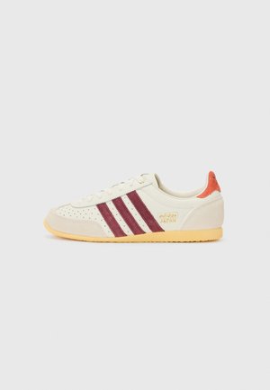 White and beige sneaker with maroon stripes, perforated detailing, and orange accent at heel. Features a textured rubber sole.