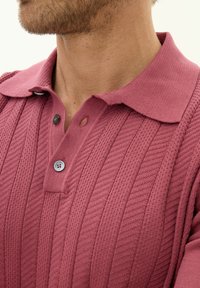 Polo shirt lavorato a maglia di colore rosa con un motivo a trama. Presenta un colletto, due bottoni e un piccolo dettaglio con asola.