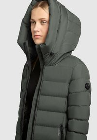 Hallrohelise värvusega polsterdatud jope, millel on suur kapuuts, esi lukuga ja küljetaskud. Omaduseks on tikitud muster ja varrukal asuv ümmargune must logo.