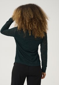 Långärmad topp i mörkgrön spets, med ett strukturerat blommönster och en figurnära design, kombinerad med svarta byxor.