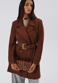 Cappotto trench marrone con vita aderente, design doppiopetto, grandi rever e accessori metallici dorati. Tiene una borsa marrone con dettagli in metallo.