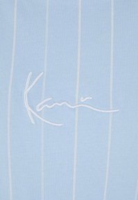 Hellblau geripptes Gewebe mit weißen vertikalen Streifen und gesticktem Signaturdetail in einem kontrastierenden Material. Lässiges und figurbetontes Design.