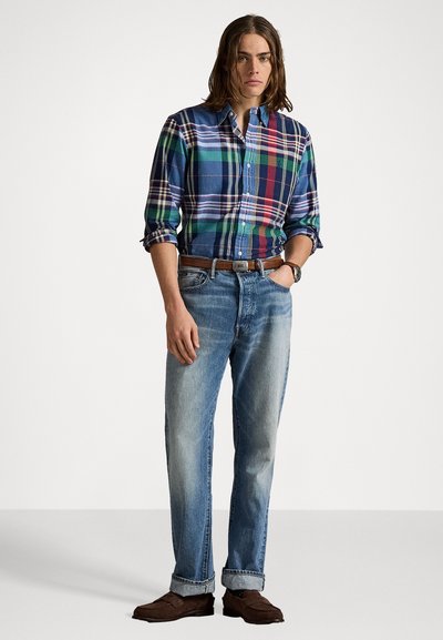 Mėlynos, žalios ir raudonos languotos marškiniai su mygtukų apykakle, dėvimi su šviesiai mėlynomis džinsais ir rudomis loafer bateliais. Paprastas rusvas diržas.