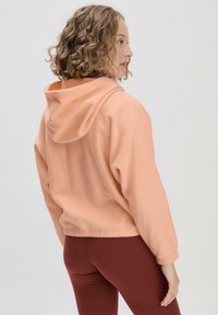 Pfirsichfarbener, verkürzter Hoodie mit Kapuze und Kordelzug, geripptem Saum und elastischen Bündchen, kombiniert mit weinroten Leggings. Weicher Materialtextur.