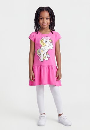 Ung pige står iført en pink kjole med et vingepony-grafik, hvide leggings og hvide sneakers mod en ensfarvet lys baggrund.