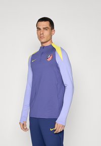 Langærmet atletisk top, lilla krop med lyse lilla ærmer, gule detaljer, Nike-logo og Atlético Madrid-våbenskjold. Lynlås-collet.