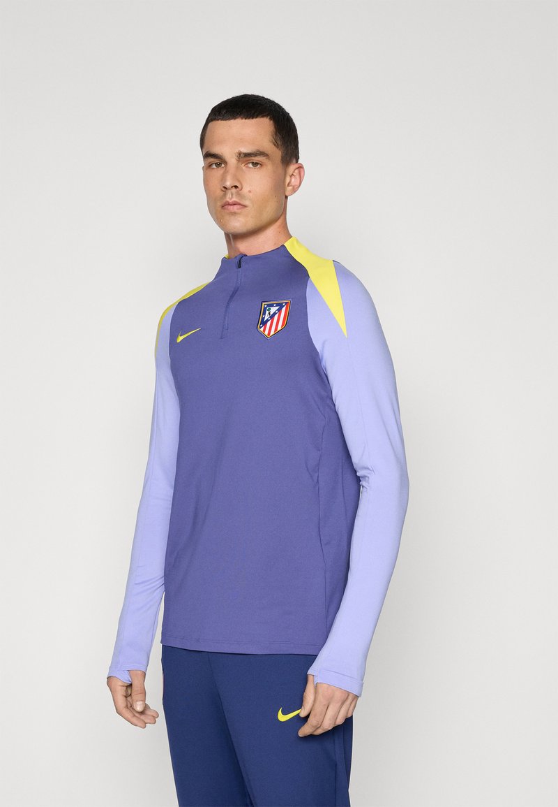Langærmet atletisk top, lilla krop med lyse lilla ærmer, gule detaljer, Nike-logo og Atlético Madrid-våbenskjold. Lynlås-collet.