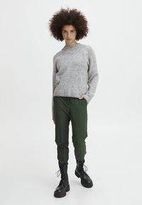 Grå strikket sweater med rund hals, grønne bukser og sorte snørebåndsstøvler med kraftige såler. Model står oprejst mod en lys baggrund.