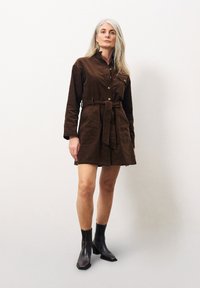 Braunes Cordshirtkleid mit langen Ärmeln, Knopfleiste und tailliertem Bund. Kombiniert mit dunklen Knöchelstiefeln. Strukturierter und maßgeschneiderter Stil.