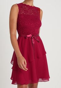 Robe rouge avec un corsage en dentelle, design sans manches et jupe en chiffon à plusieurs couches. Comprend une ceinture en ruban de satin à la taille.
