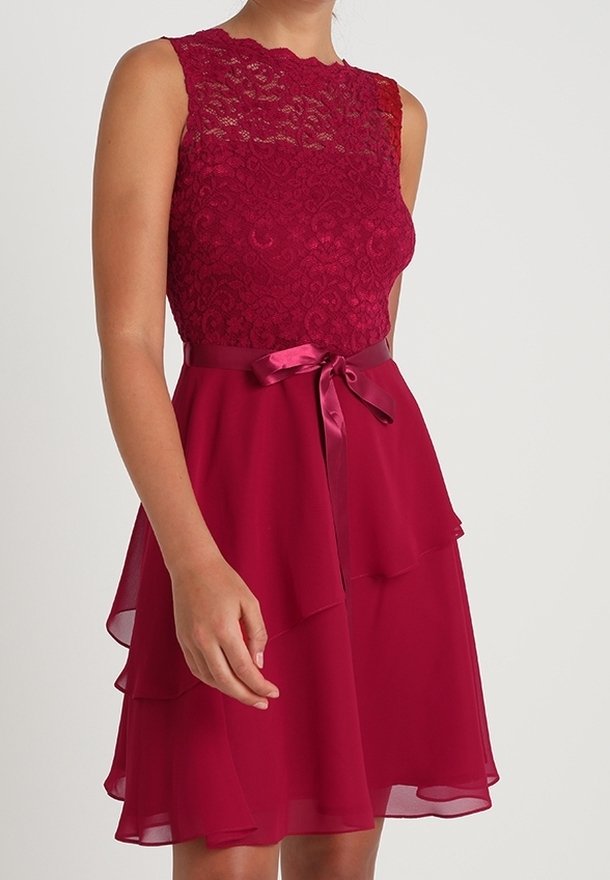 Robe rouge avec un corsage en dentelle, design sans manches et jupe en chiffon à plusieurs couches. Comprend une ceinture en ruban de satin à la taille.