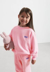 Felpa in pile rosa con polsini a coste, decorata con il logo blu "AMERICAN VINTAGE", abbinata a pantaloni della tuta coordinati, tenuti insieme a un peluche.