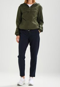 Olivgrön lätt jacka med dragkedja framtill, ihop med navyblå ränderade byxor och vita sportskor. Avslappnad design, elastiska ärmslut.