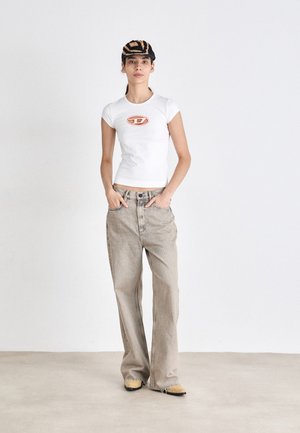 T-shirt with a circular orange logo on a white fitted base, paired with light beige wide-leg jeans and yellow shoes. Black and gold hat.