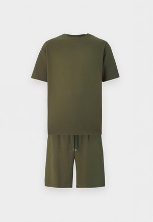 Άνδρας που φοράει φαρδιά ελιά πράσινη μπλούζα και ταιριαστά σορτς με καφέ παπούτσια χωρίς κορδόνια, στέκεται με τα χέρια στις τσέπες μπροστά σε έναν λευκό τοίχο.