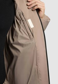 Lette beige jakke med en glat tekstur og lynlås. Indvendigt har den et lille stofmærke nær sømmen.