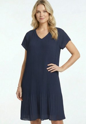 Femme aux longs cheveux blonds porte une robe bleu marine plissée à manches courtes et col en V, debout avec une main sur la hanche et l'autre détendue.