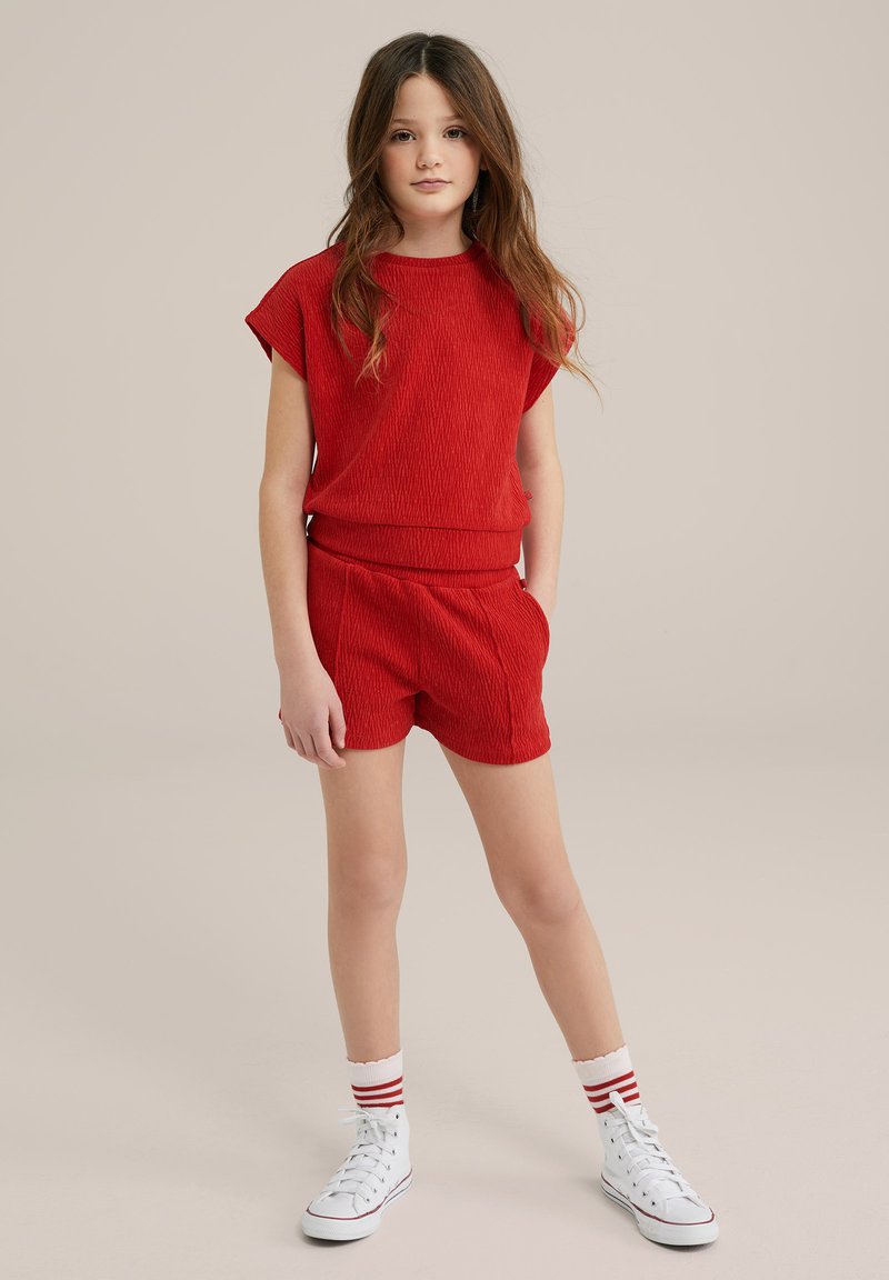 Červený textúrovaný pletený top s krátkym rukávom a zodpovedajúce šortky, doplnené o biele vysoké tenisky a podkolienky s červenými pruhmi.