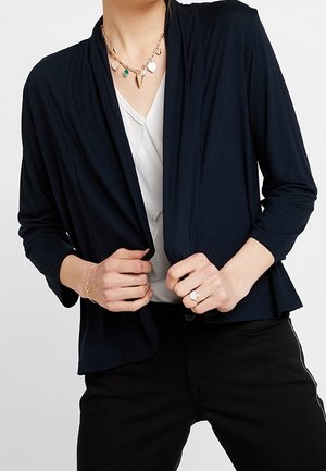 Personne portant un cardigan noir ouvert devant sur un haut blanc, un pantalon noir, un collier pendentif en or, un bracelet en or et une bague à la main droite.