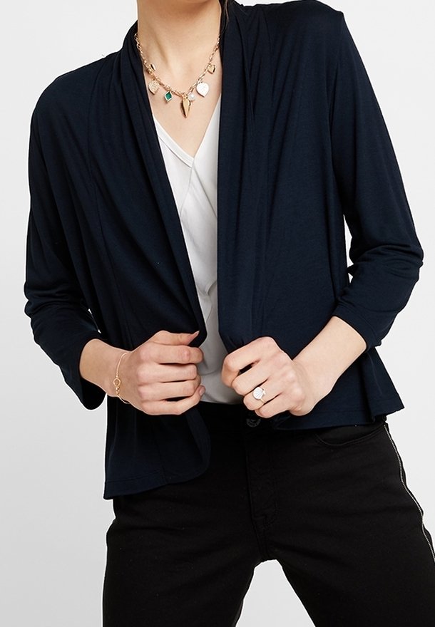 Personne portant un cardigan noir ouvert devant sur un haut blanc, un pantalon noir, un collier pendentif en or, un bracelet en or et une bague à la main droite.