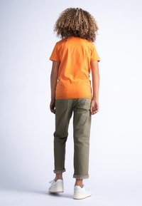 T-shirt in cotone arancione, modello aderente, abbinata a chinos verde oliva arrotolati e sneakers bianche, vista da dietro.