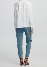 Blouse blanche à manches longues, coupe ample, associée à un jean tapered bleu clair et des sandales à talons bruns. Tissu lisse avec un subtile éclat.