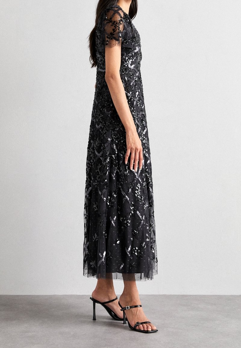 Femme portant une robe noire à sequins longue jusqu'aux chevilles avec des manches courtes transparentes et des sandales noires à talons hauts, debout sur un sol gris.