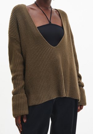 Personne portant un pull en maille marron ample avec un profond col en V par-dessus un top dos nu noir et un pantalon noir sur un fond blanc uni.