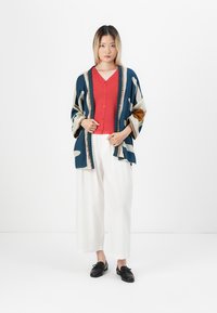 Veste de style kimono multicolore présentant des formes abstraites en bleu, crème et marron. Associée à un haut rouge à boutons et un pantalon large blanc.