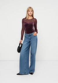 Langemouwige bordeauxrode top van doorschijnende stof met een sweetheart-halslijn, gecombineerd met wijde blauwe jeans. Een zwarte handtas voegt contrast toe.