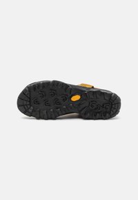 Fekete gumitalp mély barázdákkal és Vibram logóval. A saroknál sárga kiegészítővel a részletek érdekében.