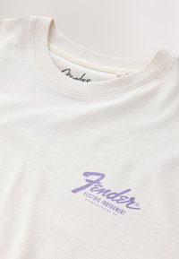 Cream cotton t-shirt featuring a purple "Fender" logo with "ELECTRIC INSTRUMENT" text below; smooth texture and classic crew neck design.