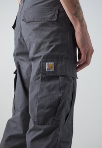 Graue Cargo-Hosen aus leichtem Stoff, mit Vorder- und Hintertaschen sowie einem Markenpatch am Oberschenkel. Strukturierte Oberfläche mit Nähten.