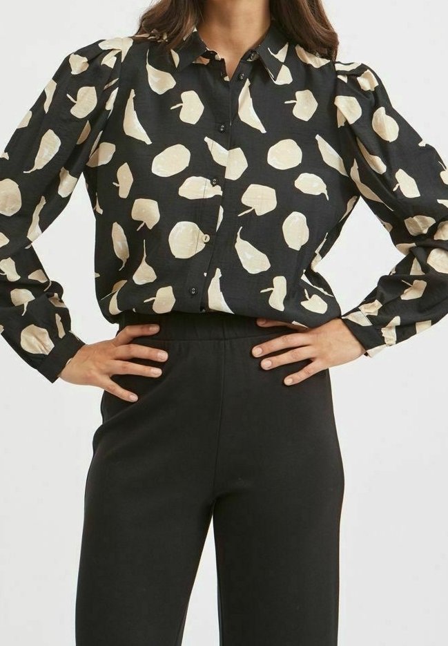 Femme portant une chemise noire boutonnée avec des taches abstraites beiges et des manches longues, associée à un pantalon noir taille haute, mains sur les hanches.