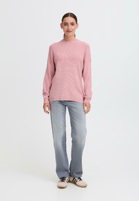 Light pink ribbed knit sweater with long sleeves, crew neck, paired with faded blue straight-leg jeans and beige sneakers with black stripes.