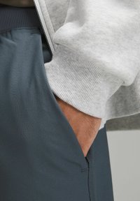 Szara hoodie na zamek błyskawiczny z prążkowanymi mankietami i ciemnoniebieskimi spodniami sportowymi. Ręka w kieszeni ukazuje gładki, teksturowany materiał.