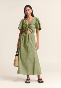 Olivno zelena dvodelna obleka z kratko, zloščeno zgornjo majico in dolgo, rahlo valovito krilo. Model drži pleteno torbo; look dopolnjujejo ravne črne sandale.