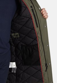 Olivinės žalos spalvos išorinis švarkas su juodu užpildo pamušalu, turintis raudoną apvadą, užtrauktuką turintį kišenę ir tinklinę saugojimo kišenę viduje.