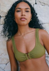 Grüner strukturierter Bikini-Oberteil mit Rundhalsausschnitt, ausgestattet mit einer Front-Drehung und Metallbeschlägen. Das Model hat welliges schwarzes Haar.