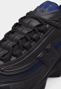 Μαύρο και μπλε παπούτσι New Balance 1000 με χοντρές κορδόνια και έντονο λογότυπο "N" στο πλάι και στη γλώσσα.