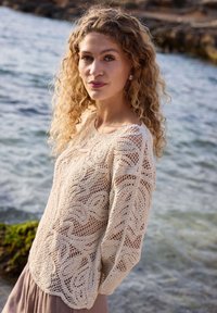 Femme aux cheveux blonds bouclés portant un pull beige en crochet, debout près d'une côte rocheuse avec une mer calme en arrière-plan.