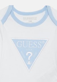 Lühikeste varrukatega valge bodi helesinise ääristusega, millel on tikitud sinine kolmnurkne logo koos tekstiga "GUESS" ja küsimärgi kujundusega.