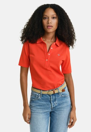 Polo rosso con maniche corte, collo a tre bottoni e dettagli del logo ricamato. Indossato con jeans blu e una cintura in tessuto color tan.
