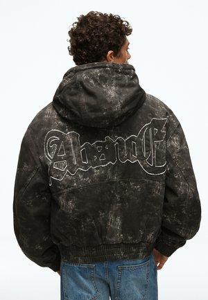 Personne aux cheveux bouclés portant une veste à capuche noire et grise mouchetée avec le mot "Absent" brodé dans le dos, associée à un jean bleu.